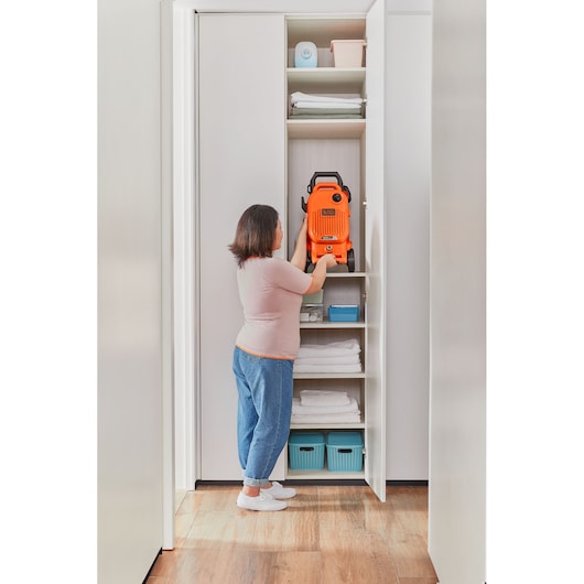 Photo of a woman storing an orange BLACK+DECKER BEPW1700 pressure washer on a shelf in a white closet.