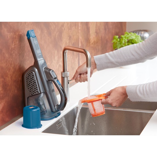 BLACKDECKER BHHV320B handheld vacuum being cleaned, filter rinsed under faucet in kitchen sink, side view.