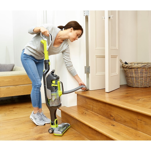 A person using a BLACK+DECKER upright vacuum cleaner with a hose attachment to clean the edge of wooden stairs near a doorway. A wicker basket is visible in the background.
