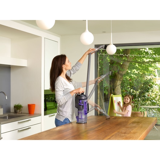 A person using a BLACK+DECKER vacuum cleaner with a long hose attachment to clean a pendant light in a modern kitchen. Another child is drawing at an easel near a window with a garden view.