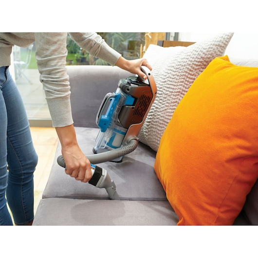 BLACKDECKER CUA525BH vacuum cleaner used on grey sofa with orange cushion in home setting.