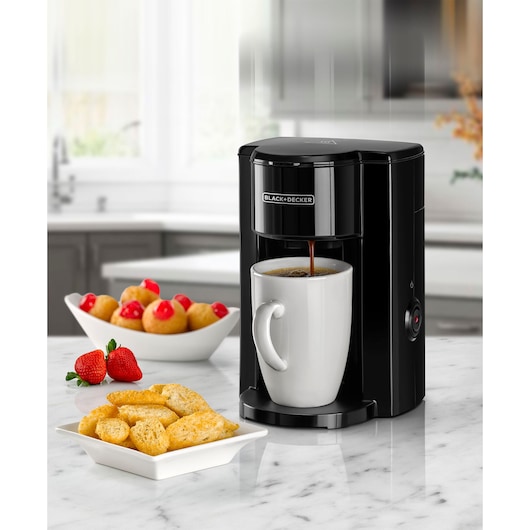 BLACK+DECKER DCM25N coffee maker brewing coffee into a white mug on a kitchen counter with pastries and strawberries.