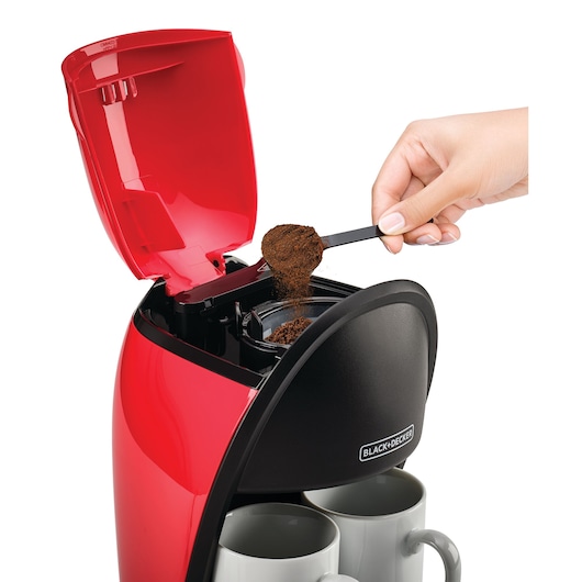 BLACKDECKER DCM48 red coffee maker front angle with person adding ground coffee to filter, two mugs below.