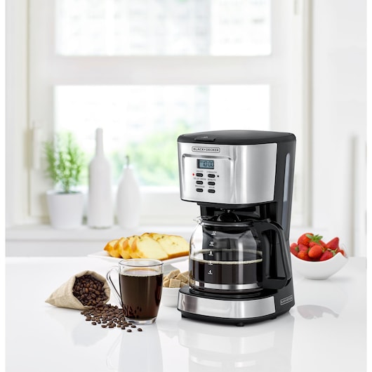A BLACK+DECKER drip coffee maker with a glass carafe is placed on a kitchen counter. Nearby, there is a cup of freshly brewed coffee, spilled coffee beans, bread slices on a plate, and a bowl of strawberries. The kitchen is bright with a large window in the background.