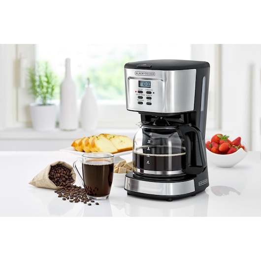 A BLACK+DECKER drip coffee maker with a digital display sits on a kitchen counter. In front of it, there is a glass mug of coffee, some coffee beans, sugar cubes, a plate of sliced cake, and a bowl of strawberries.