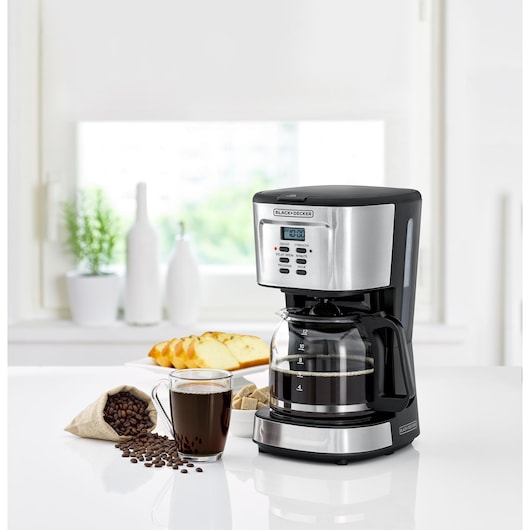A BLACK+DECKER drip coffee maker with a digital control panel displays on a kitchen counter. Next to it, there is a glass mug filled with coffee, a small burlap sack of coffee beans, and slices of bread on a plate. The background shows some decorative bottles and a green plant by a window.