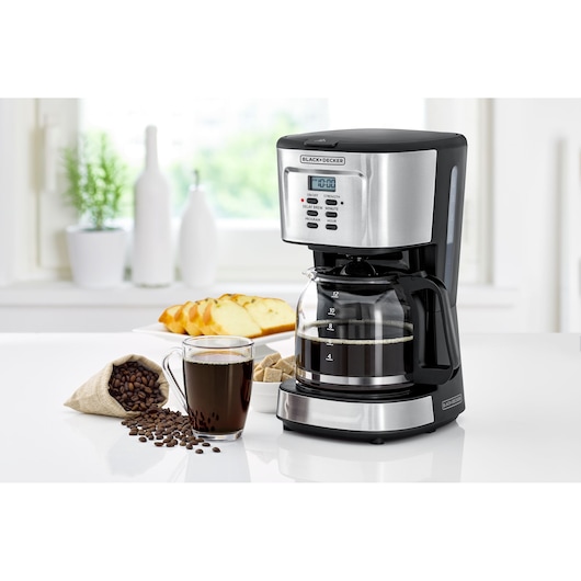 A BLACK+DECKER drip coffee maker with digital display sits on a kitchen counter. Next to it is a clear glass mug filled with coffee, a small burlap bag of coffee beans, sugar cubes, and a plate with sliced bread. The background shows white vases and plants.