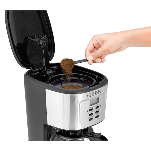 A hand is adding ground coffee to the open filter compartment of a BLACK+DECKER coffee maker.