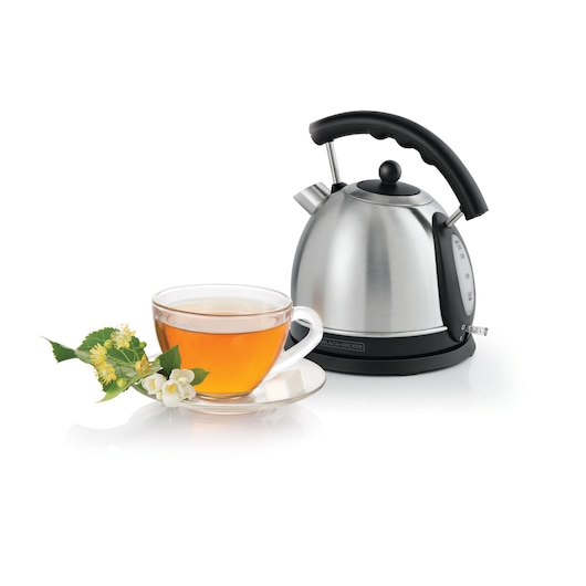 A stainless steel electric kettle with a black handle next to a glass cup of tea garnished with flowers and a sugar cube on a saucer.