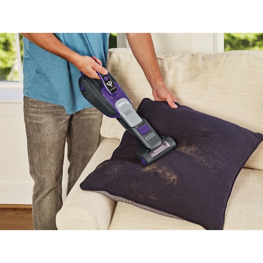 BLACKDECKER DVJ325BFSP handheld vacuum cleaning pet hair from a dark pillow on a beige sofa.