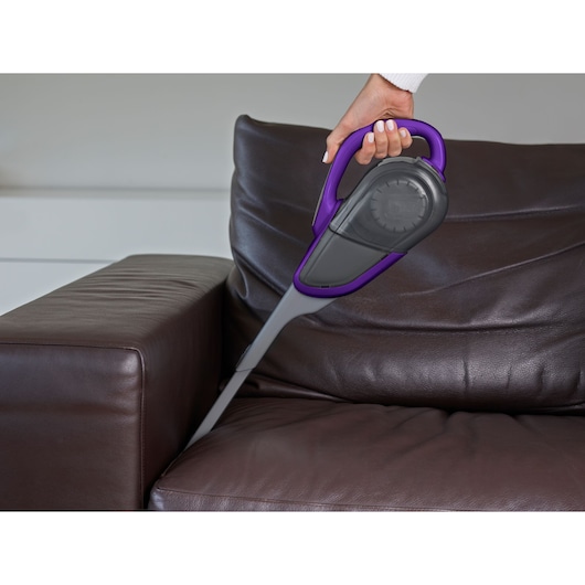 BLACKDECKER DVJ325BFSP handheld vacuum with purple accents cleaning a brown leather sofa.