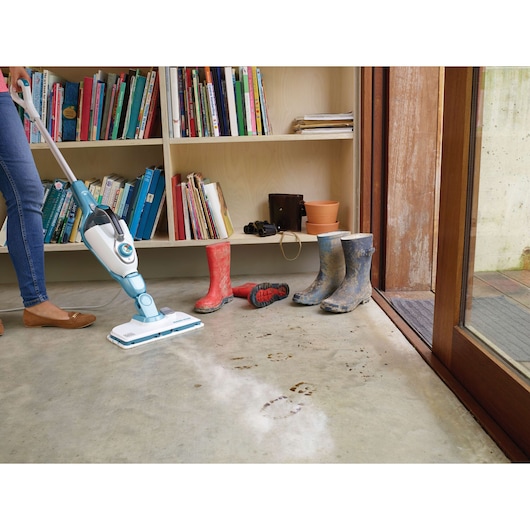 BLACKDECKER FSMH13151SM steam mop cleaning muddy footprints on concrete floor near boots and bookshelf.