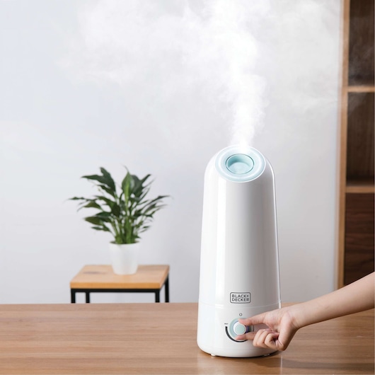 Photo of BLACK+DECKER HM5000 humidifier releasing mist on a tabletop, hand adjusting control knob.