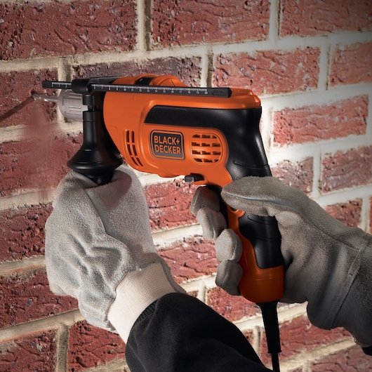 A person wearing work gloves is using a BLACK+DECKER electric drill to drill into a brick wall.