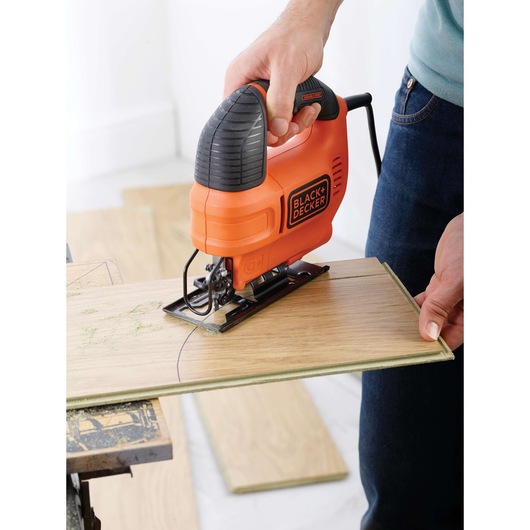 A person using an orange BLACK+DECKER jigsaw to cut a piece of wood.