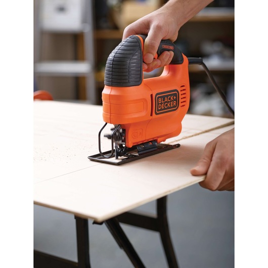 A person uses a BLACK+DECKER orange jigsaw to cut a piece of wood on a workbench.