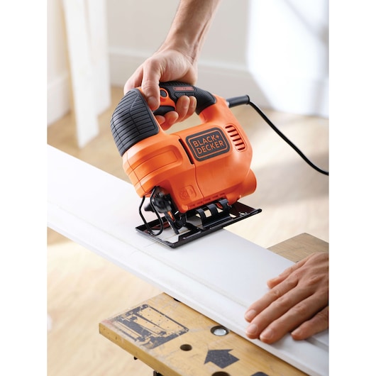 A person using a BLACK+DECKER KS701EK electric jigsaw to cut a piece of wood, with one hand guiding the tool and the other holding the wood steady on a workbench.
