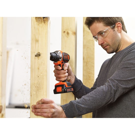 A person using a BLACK+DECKER cordless drill to make a hole in a wooden beam. The person's face is blurred for privacy, and wood shavings are visible as the drill operates.