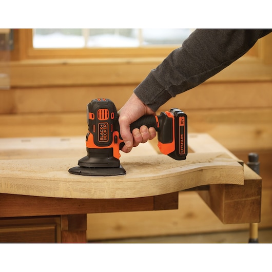A person using a BLACK+DECKER cordless electric sander to smooth the surface of a wooden plank in a workshop.