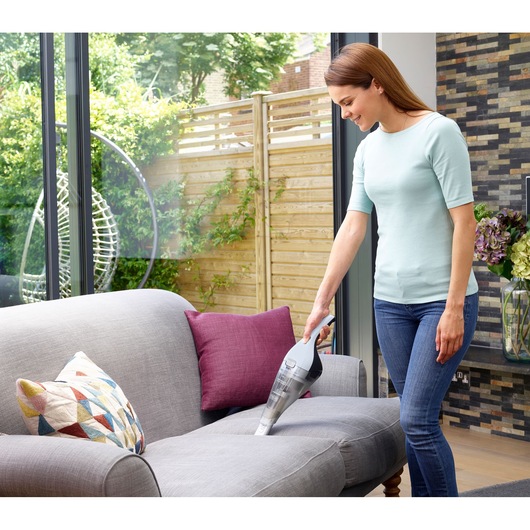 BLACKDECKER NVC215W handheld vacuum cleaning sofa upholstery with grey and maroon cushions in living room.