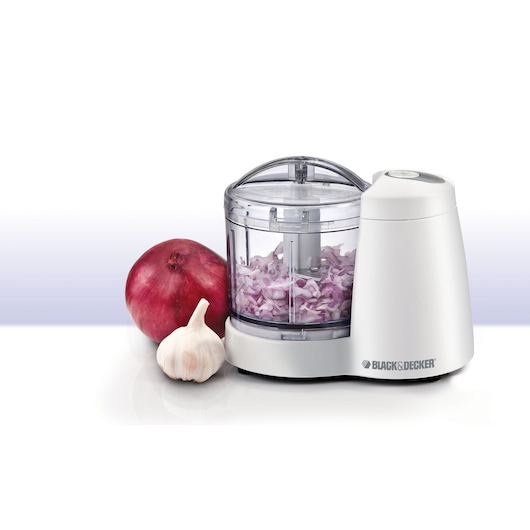 A white BLACK+DECKER mini food chopper with chopped onions inside, displayed next to a whole red onion and a garlic bulb on a white surface.