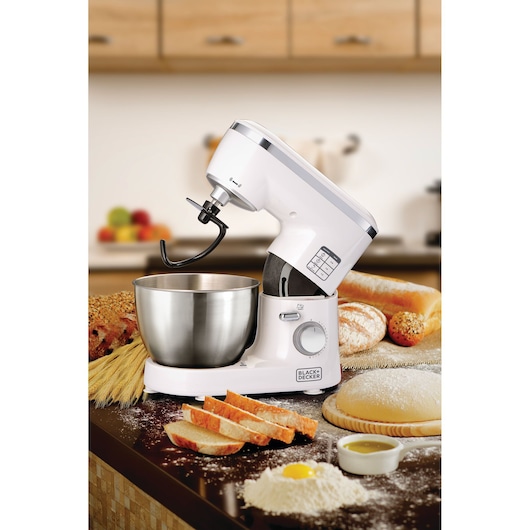 A BLACK+DECKER stand mixer, model SM1000, in white color with a stainless steel mixing bowl, is placed on a kitchen counter surrounded by bread, flour, an egg, and baking ingredients. The mixer features a dough hook attachment, with a background showing a modern kitchen setting.