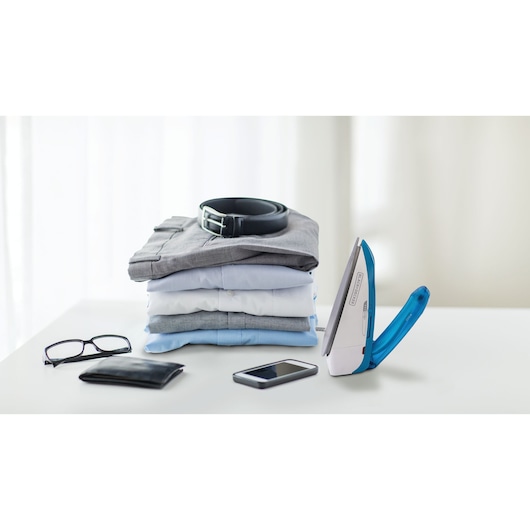 BLACKDECKER TI250 travel iron blue and white on table beside folded shirts, belt, glasses, wallet, and phone, side angle.
