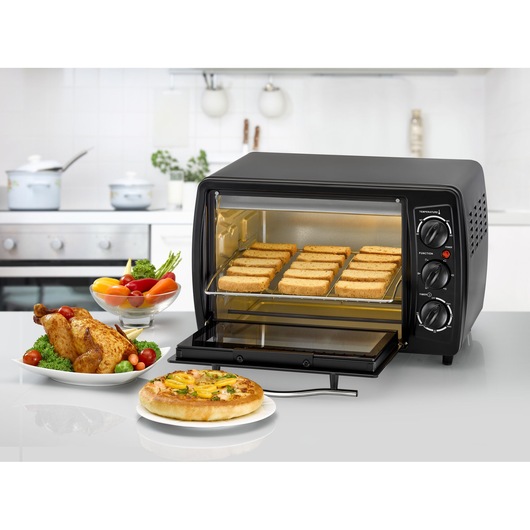A BLACK+DECKER countertop toaster oven with an open door, filled with slices of toast. In the foreground, there is a roasted chicken on a plate with vegetables, a bowl of assorted fresh vegetables, and a pizza on a plate, all placed on a kitchen counter. The kitchen is modern with white cabinetry and a stove in the background.