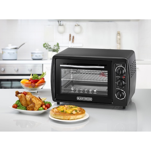A BLACK+DECKER countertop electric oven with three control knobs, placed in a modern kitchen. In front of the oven are a plate of roasted chicken with vegetables, a bowl of fresh vegetables, and a small pizza on a plate.
