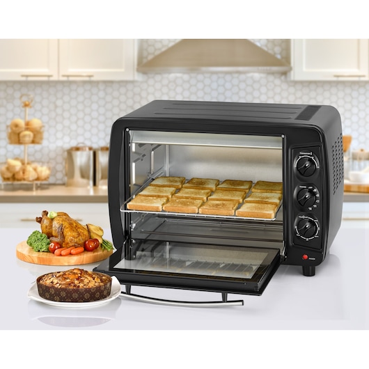 A BLACK+DECKER countertop oven with a tray of toasted bread slices inside. The oven has three control dials on the right side and its door is open. In front of the oven, there is a roasted chicken with vegetables on a wooden board and a round cake on a plate. The scene is set in a modern kitchen.