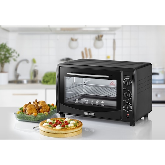 A black BLACK+DECKER countertop oven with three control knobs and a transparent door is seen on a white kitchen counter. Next to the oven are a whole roasted chicken in a glass dish with vegetables and a pizza on a wooden board. The kitchen background features white tiles and utensils.