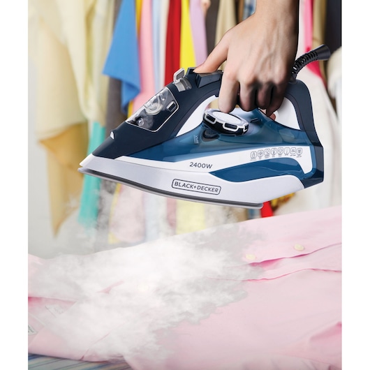 BLACK+DECKER X2150 steam iron in use on pink shirt, steam visible, clothes hanging in background.