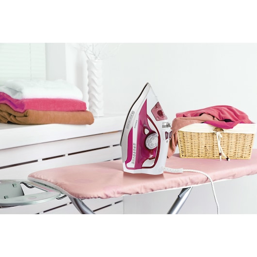 BLACKDECKER X2400 pink and white steam iron on ironing board with folded towels and laundry basket in background.